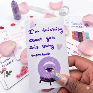 Könnte beinhalten: Eine weiße Karte mit der handgeschriebenen Nachricht "I'm thinking about you this very moment" in lila Tinte. Auf der Karte befindet sich ein lila Augenaufkleber. Weitere Karten, Kristalle und Rosenblätter sind im Hintergrund.