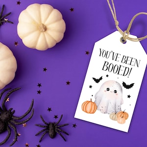 May include: A Halloween-themed image with a white tag saying "YOU'VE BEEN BOOED!" and a ghost illustration. The tag is tied with twine, surrounded by white pumpkins, black spiders, and star-shaped confetti on a purple background.