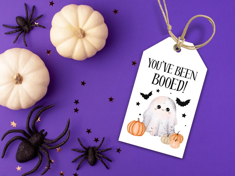 May include: A Halloween-themed image with a white tag saying "YOU'VE BEEN BOOED!" and a ghost illustration. The tag is tied with twine, surrounded by white pumpkins, black spiders, and star-shaped confetti on a purple background.