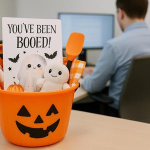 May include: An orange Halloween bucket with a jack-o'-lantern face design, filled with a card that reads "YOU'VE BEEN BOOED!" and a plush ghost. Also includes a small pumpkin, a spatula, and a checkered cloth. A person is blurred in the background.