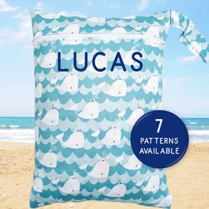 May include: A blue and white patterned wet bag featuring a whale design. The bag has a white zipper and a handle. The name "LUCAS" is printed on the bag. A circular graphic states "7 PATTERNS AVAILABLE".