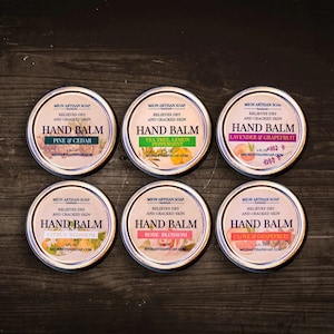 May include: Six round metal tins with lids, each featuring a different hand balm scent. The tins are labeled with the scent and the brand name "Mion Artisan Soap".