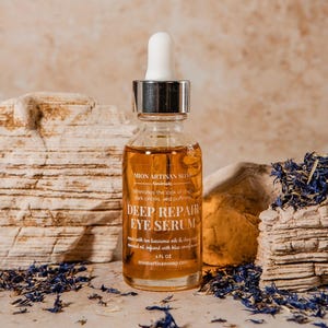 May include: A clear glass bottle of Deep Repair Eye Serum with a white dropper, set against a neutral background with dried blue flowers. The label reads "MION ARTISAN SOAP" and "DEEP REPAIR EYE SERUM".