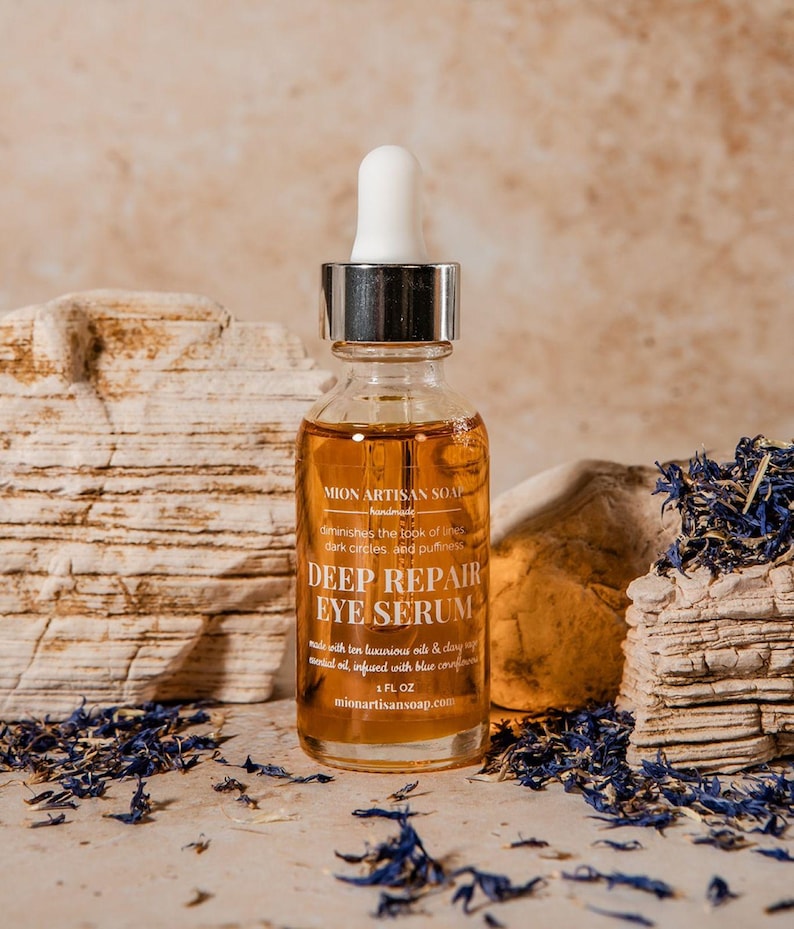 May include: A clear glass bottle of Deep Repair Eye Serum with a white dropper, set against a neutral background with dried blue flowers. The label reads "MION ARTISAN SOAP" and "DEEP REPAIR EYE SERUM".
