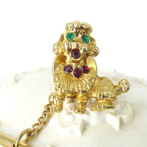 May include: Gold-tone tie tack featuring a poodle with red and green rhinestones. The poodle is sitting with its head tilted and has a chain attached to the back.