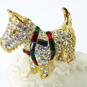 May include: A gold-tone brooch in the shape of a Scottish terrier, adorned with clear rhinestones and colorful enamel accents. The dog is wearing a red, green, and blue tartan ribbon.