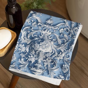 May include: A blue and white decorative towel with a floral and scroll design. The towel is folded on a small, dark table. A black bottle and a wooden soap dish are also on the table. The towel's design features a central floral motif.