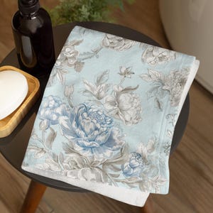 May include: A folded pale blue bath towel with a floral design of large blue and grey peonies. The towel is on a small, dark table. A dark brown bottle and a bar of soap are also visible.