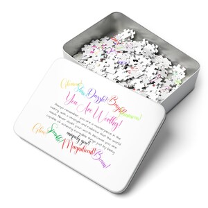 May include: A white puzzle tin with the lid open, revealing a collection of puzzle pieces. The lid features colorful text that reads "You Are Worthy!" and inspirational phrases. The tin is silver.