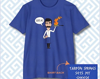 May include: A royal blue t-shirt featuring a cartoon character with a mustache holding a flaming pan with the Greek text ΩΠΑ! in a speech bubble. The lower portion includes the text SHIRT BACK and TARPON SPRINGS SETS MY CHEESE ON FIRE!