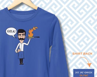 May include: A royal blue long-sleeve shirt featuring a cartoon illustration of a person with a mustache holding a flaming pan. A speech bubble contains the Greek text ΩΠΑ!. The text SET MY CHEESE ON FIRE! is printed in a separate blue box.