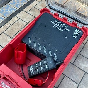 Puede incluir: Una caja de almacenamiento roja Milwaukee Packout que contiene un comprobador de luces de remolque inalámbrico negro. El comprobador tiene etiquetas para varias funciones de luz y un pequeño mando a distancia negro. También se ven un soporte rojo y un conector rojo.