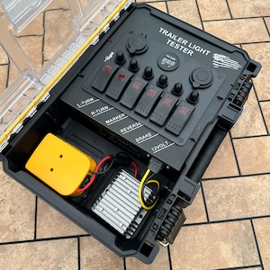 May include: A black trailer light tester with a clear and yellow lid. The tester has multiple switches labeled L-TURN, R-TURN, MARKER, REVERSE, and BRAKE. It also features a digital voltage display and the text "TRAILER LIGHT TESTER".