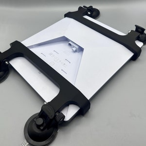 Könnte beinhalten: Ein schwarzer Tablet-Halter mit Saugnäpfen an der Unterseite. Der Halter ist so konzipiert, dass er ein Tablet sicher an seinem Platz hält. Das Tablet ist nicht im Lieferumfang enthalten.