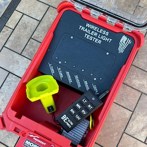Puede incluir: Una caja Packout roja que contiene un comprobador de luces de remolque inalámbrico. La caja contiene un panel de control negro con luces indicadoras, un asa amarilla y un mando a distancia negro. El comprobador es para diagnósticos.