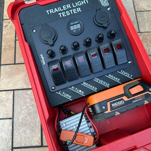 May include: Red trailer light tester with black control panel. Features include switches labeled L-TURN, R-TURN, MARKER, REVERSE, and BRAKE. Includes a digital voltage display and a Ridgid battery.