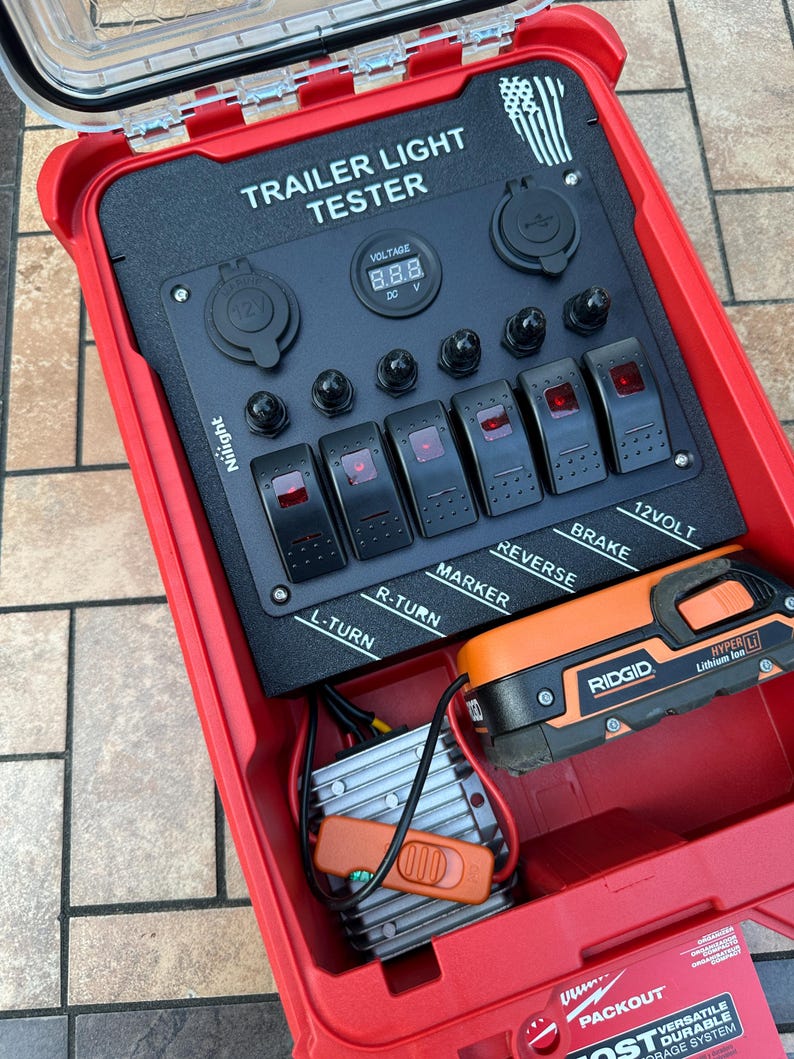 May include: Red trailer light tester with black control panel. Features include switches labeled L-TURN, R-TURN, MARKER, REVERSE, and BRAKE. Includes a digital voltage display and a Ridgid battery.