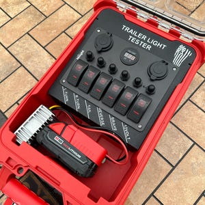 以下が含まれることがあります： 複数のスイッチとデジタルディスプレイが付いた「トレーラーライトテスター」というラベルの付いた黒いパネルが付いた赤いツールボックス。ツールボックスが開いており、「M18 REDLITHIUM」というラベルの付いた黒と赤のバッテリーが入っています。