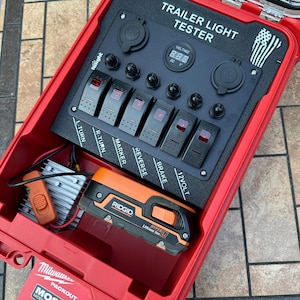 May include: A red Milwaukee Packout case containing a trailer light tester. The tester has multiple switches labeled L-TURN, R-TURN, MARKER, REVERSE, BRAKE, and 12VOLT. A digital display shows voltage. A Ridgid Hyper Lithium Ion battery is also inside.
