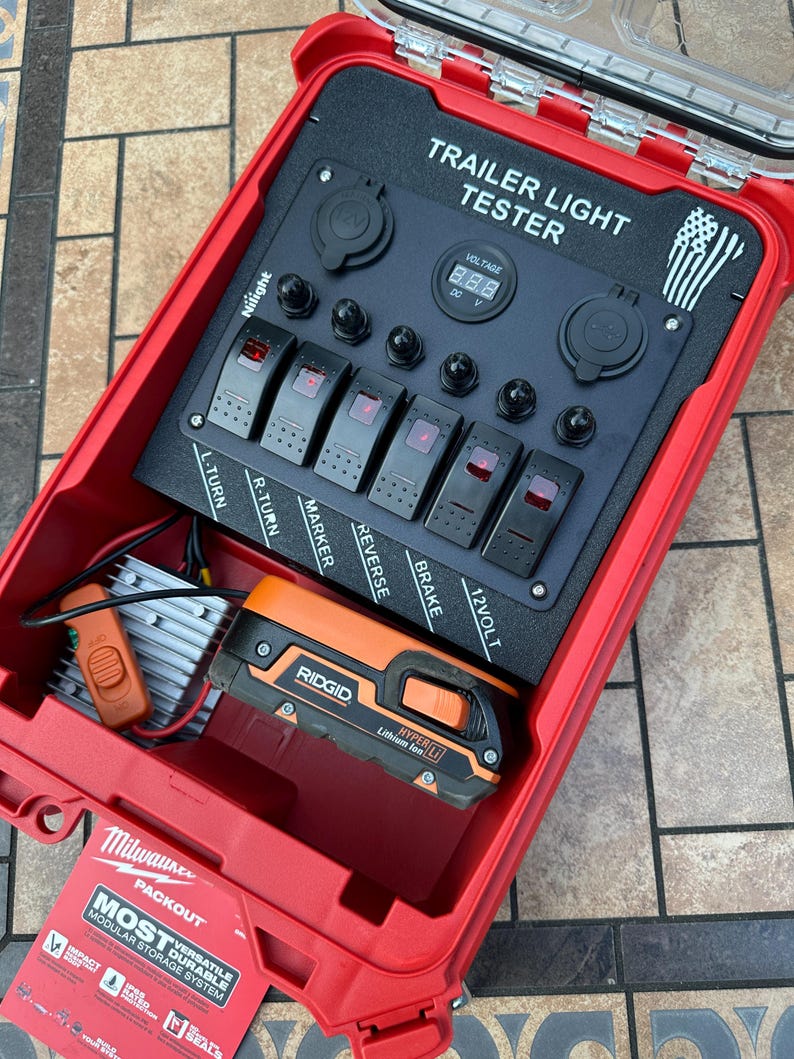 May include: A red Milwaukee Packout case containing a trailer light tester. The tester has multiple switches labeled L-TURN, R-TURN, MARKER, REVERSE, BRAKE, and 12VOLT. A digital display shows voltage. A Ridgid Hyper Lithium Ion battery is also inside.