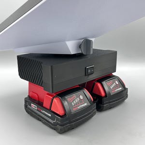 Op de afbeelding: Twee rode Milwaukee M18 RedLithium-batterijen zijn verbonden met een zwarte powerbank met een wit lampje en een zwarte knop. De powerbank heeft een zwarte aan/uit-schakelaar.
