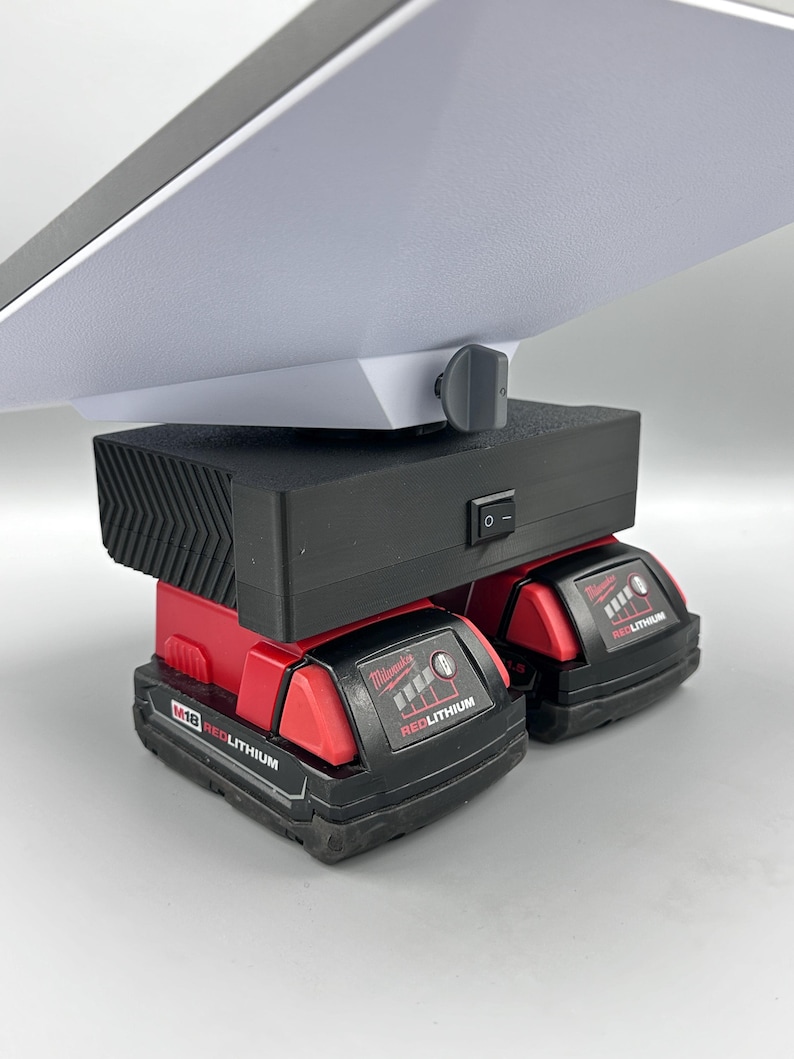 Op de afbeelding: Twee rode Milwaukee M18 RedLithium-batterijen zijn verbonden met een zwarte powerbank met een wit lampje en een zwarte knop. De powerbank heeft een zwarte aan/uit-schakelaar.
