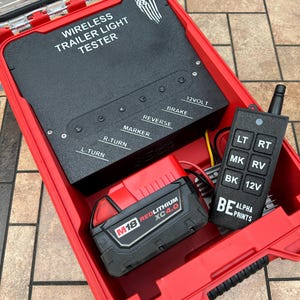 Pu&ograve; includere: Una custodia rossa contenente un tester per luci per rimorchi wireless nero con la scritta "WIRELESS TRAILER LIGHT TESTER". Include un telecomando nero e un pacco batteria rosso e nero. La custodia &egrave; aperta, rivelando i componenti.