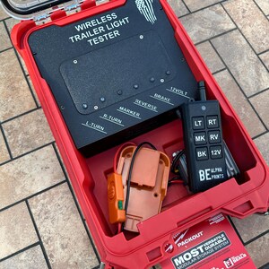 May include: A red Packout case with a clear lid houses a wireless trailer light tester. The black tester has labels for brake, reverse, and turn signals. A remote control and orange components are also inside. The case has the text "MOST VERSATILE DURABLE MODULAR STORAGE SYSTEM."