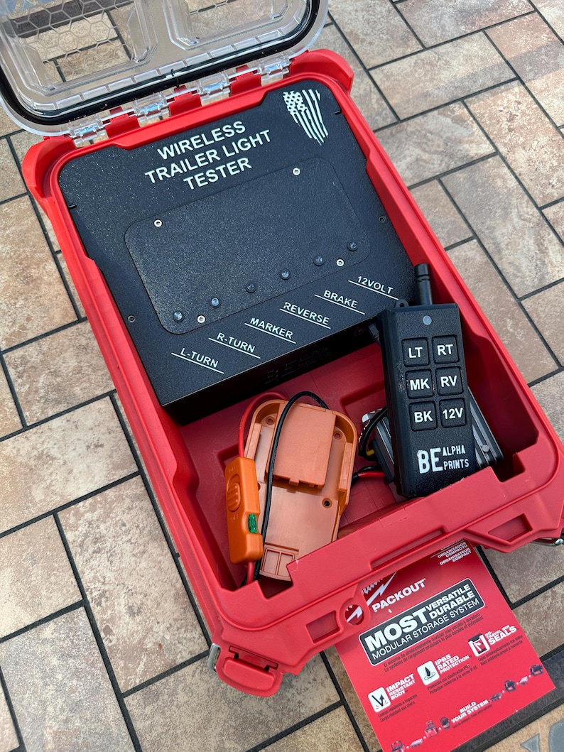 May include: A red Packout case with a clear lid houses a wireless trailer light tester. The black tester has labels for brake, reverse, and turn signals. A remote control and orange components are also inside. The case has the text "MOST VERSATILE DURABLE MODULAR STORAGE SYSTEM."