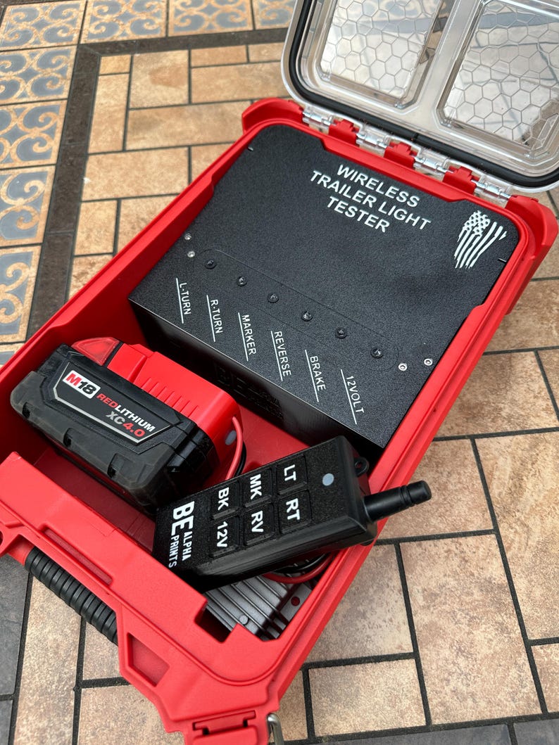 Pu&ograve; includere: Una custodia rossa, aperta e impermeabile contenente un tester per luci per rimorchi wireless nero, una batteria rossa e nera e un telecomando nero. Il tester ha il testo "WIRELESS TRAILER LIGHT TESTER". Il telecomando ha il testo "LT RT MK RV BK 12V".