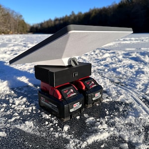 May include: Two red and black M18 XC6.0 lithium-ion batteries are stacked on top of a black base with a white and gray rectangular panel on top. The panel is angled upwards and appears to be a light source. The batteries and panel are sitting on a snowy surface.