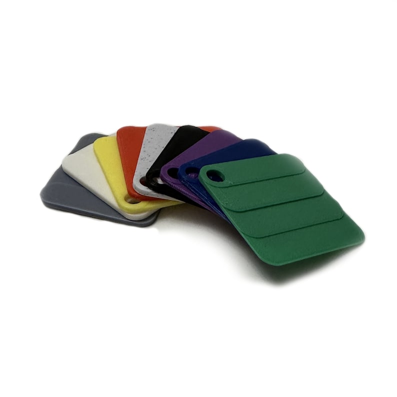 May include: A set of eight small, square plastic tiles in various colours, including grey, white, yellow, red, speckled grey, black, purple, and blue. The tiles are arranged in a fan shape, with the green tile at the bottom.