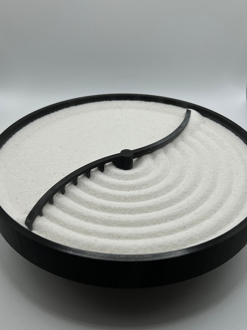 May include: A black circular tray with a white sand base and a black plastic divider creating a yin yang design. The sand is textured and has a series of concentric circles on one side.