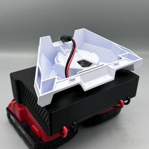 Op de afbeelding: Een witte plastic oplaadbasis met een zwart stroomsnoer en rode en zwarte draden. De basis is open en toont een zwart cirkelvormig gebied waar een batterij zou worden geplaatst. De basis staat op een zwart en rood batterijpakket.