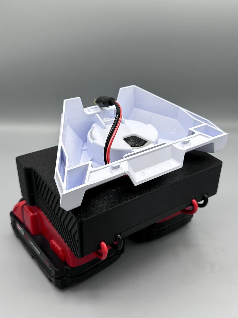 Op de afbeelding: Een witte plastic oplaadbasis met een zwart stroomsnoer en rode en zwarte draden. De basis is open en toont een zwart cirkelvormig gebied waar een batterij zou worden geplaatst. De basis staat op een zwart en rood batterijpakket.