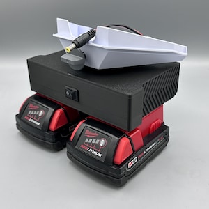 Op de afbeelding: Een wit oplaadstation met een zwart stroomsnoer en een zwarte aan/uit-schakelaar. Het oplaadstation staat op twee rode en zwarte Milwaukee M18 RedLithium-accu's.