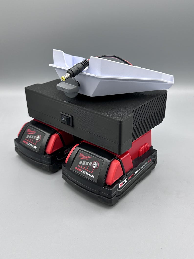 Op de afbeelding: Een wit oplaadstation met een zwart stroomsnoer en een zwarte aan/uit-schakelaar. Het oplaadstation staat op twee rode en zwarte Milwaukee M18 RedLithium-accu's.