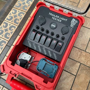Puede incluir: Un comprobador de luces de remolque rojo con un panel de control negro. El panel tiene interruptores etiquetados como "L TURN", "R TURN", "MARKER", "REVERSE", "BRAKE" y "12VOLT". También cuenta con una pantalla digital de voltaje y dos tomas de corriente. La carcasa está abierta, revelando componentes internos.