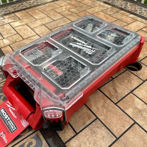 Pu&ograve; includere: Un sistema di stoccaggio modulare Milwaukee Packout rosso e trasparente. Il coperchio trasparente rivela diversi scomparti. Il lato aperto mostra un'etichetta rossa con le parole "MOST VERSATILE DURABLE MODULAR STORAGE SYSTEM".