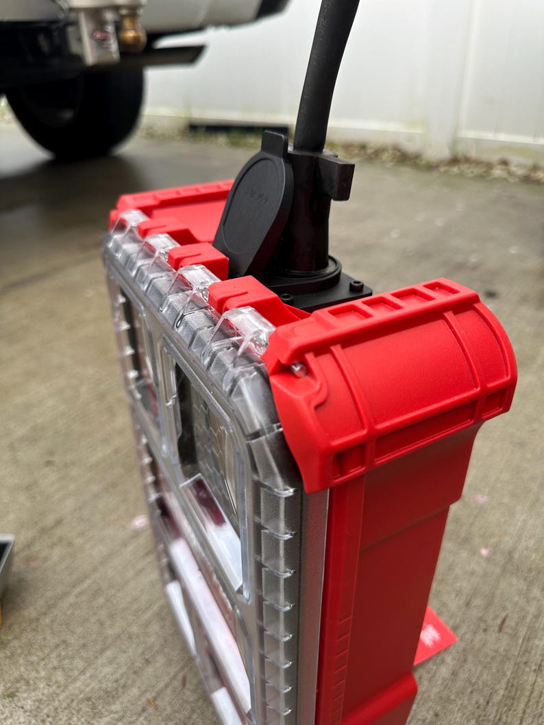 Pu&ograve; includere: Un organizer per utensili in plastica rossa e trasparente con un cavo di alimentazione e una spina neri collegati. L'organizer ha un coperchio trasparente e chiusure e dettagli rossi. L'immagine &egrave; stata scattata all'aperto.