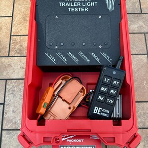 May include: A red Packout case containing a wireless trailer light tester. The tester has a black control panel with labels for lights and a remote control. The case also holds an orange adapter and a black remote control.