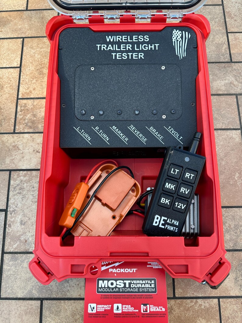 May include: A red Packout case containing a wireless trailer light tester. The tester has a black control panel with labels for lights and a remote control. The case also holds an orange adapter and a black remote control.
