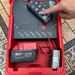 Pu&ograve; includere: Un dispositivo portatile rosso e nero con un pannello di controllo e un telecomando nero. Il telecomando ha pulsanti con le etichette "LT", "RT", "MK", "RV", "BK" e "12V". Il dispositivo contiene anche una batteria rossa e un componente argentato.