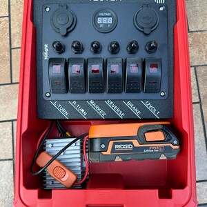 May include: A red trailer light tester with the text "TRAILER LIGHT TESTER". It has a digital voltage display, multiple switches labeled for trailer functions, and a Ridgid Hyper Lithium Ion battery.
