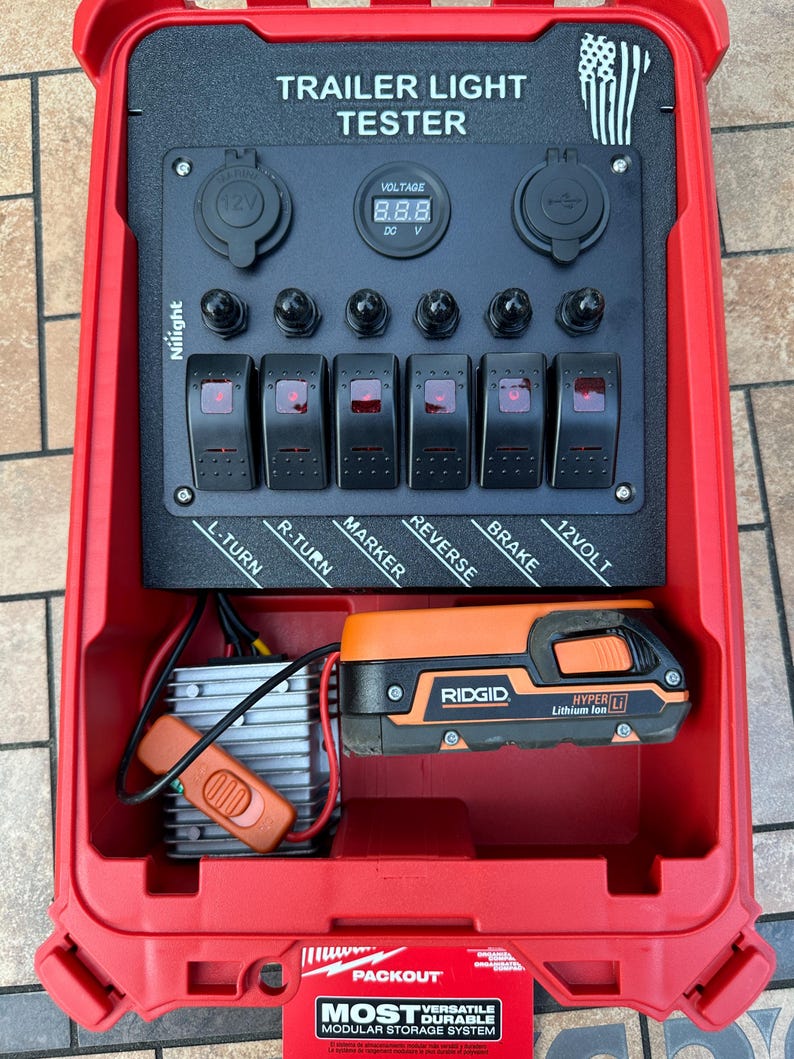 May include: A red trailer light tester with the text "TRAILER LIGHT TESTER". It has a digital voltage display, multiple switches labeled for trailer functions, and a Ridgid Hyper Lithium Ion battery.