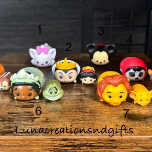 May include: A collection of colorful, round character figurines, including a white cat with a pink bow, Mickey Mouse, and Simba. The figurines are arranged on a wooden surface. The text "Lunacreationsndgifts" is at the bottom.