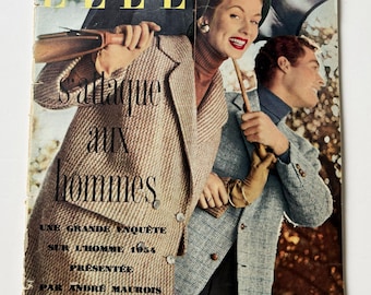 May include: A vintage October 1954 issue of Elle magazine featuring a cover with a person in a tan textured coat and another in a blue plaid blazer holding an umbrella. The text reads Elle, s'attaque aux hommes, une grande enquête sur l'homme 1954.