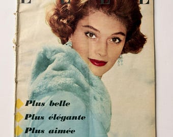 May include: A vintage Elle magazine cover from January 16, 1956, featuring a portrait of a person with brown hair wearing a light blue fur-collared coat and blue earrings. The text reads Elle, Plus belle, Plus élégante, Plus aimée, and more.