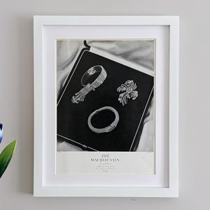 May include: A framed black and white print featuring a jewelry display with a bracelet, a brooch, and a watch inside a case. The text at the bottom reads MAUBOUSSIN, 3 Rue de Choiseul, 16 Place Vendome, PARIS. The frame is white with a white mat board.