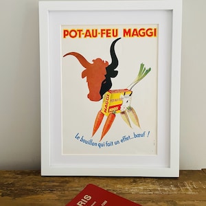 May include: Framed vintage advertisement for Maggi Pot-au-Feu, featuring a bull's head composed of vegetables. The poster has text in French, with a white frame. A red Paris guide is in the foreground.